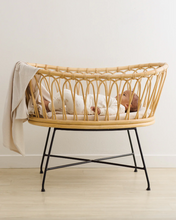 Load image into Gallery viewer, Baby sleeping in a wicker crib with a white blanket on a light wooden floor.

