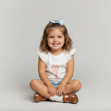 Load image into Gallery viewer, Young girl sitting on the floor wearing a white shirt with a design and blue shorts, smiling at the camera.
