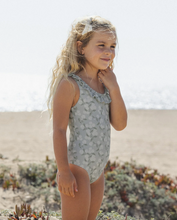 Load image into Gallery viewer, Young girl in a floral swimsuit standing on a beach with sand and plants in the background.
