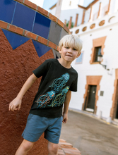 Load image into Gallery viewer, Child wearing a black t-shirt with a colorful design in front of a building.
