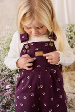 Load image into Gallery viewer, A young blonde girl wearing a white ribbed turtleneck and dark purple cherry-patterned bib overalls, looking down while touching the leather brand patch on the chest.
