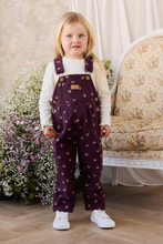 Load image into Gallery viewer, A young girl with blonde hair stands wearing a pair of dark purple cherry print overall pants over a white long-sleeve shirt and white sneakers. She is posed in front of a vintage floral armchair and a large floral arrangement.
