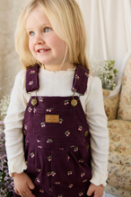 Load image into Gallery viewer, A young blonde girl wearing a white long-sleeve top and dark purple corduroy overalls with a cherry print and a tan brand patch on the chest pocket, standing in a softly lit indoor setting.
