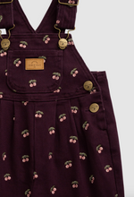 Load image into Gallery viewer, Close-up of a dark plum corduroy overalls with a cherry print, featuring gold-tone metal buttons and a tan brand patch on the chest pocket.
