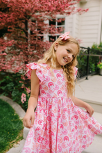 Load image into Gallery viewer, Young girl in a pink dress with horseshoe pattern standing outdoors near a building with cherry blossoms.
