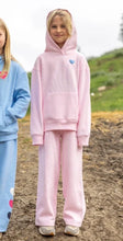 Load image into Gallery viewer, A child wearing a light pink hooded sweatshirt and matching sweatpants stands outdoors on a dirt path, with another child partially visible on the left in a blue matching set.
