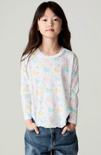 Load image into Gallery viewer, A young girl with long dark hair stands against a light gray studio background wearing a white long-sleeve top with a pastel unicorn print and blue jeans.
