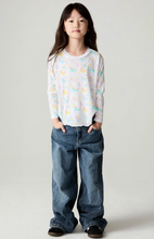 Load image into Gallery viewer, A young girl stands against a light gray studio background wearing a white long-sleeve t-shirt with a pastel unicorn print and loose blue jeans.
