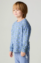 Load image into Gallery viewer, A child wearing a light blue long-sleeve shirt with a repeating pattern of green dinosaurs and white lightning bolts, paired with matching blue pants, photographed against a plain light gray studio background.
