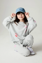 Load image into Gallery viewer, A young girl wearing a light gray sweatshirt and matching jogger pants with pink cherry graphics, a blue baseball cap, and white sneakers, posing on a light gray studio background.
