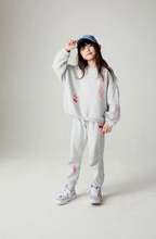 Load image into Gallery viewer, A young girl models a light gray heathered two-piece tracksuit with pink and red cherry patches, a blue baseball cap, and gray sneakers against a plain light gray studio background.
