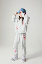 Load image into Gallery viewer, A young girl modeling a light gray heathered two-piece tracksuit with pink cherry and bow graphics, paired with a blue baseball cap and white sneakers, against a plain light gray studio background.
