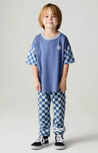 Load image into Gallery viewer, A young boy modeling a matching two-piece pajama set consisting of a blue short-sleeve top with blue-and-white checkered sleeves and matching checkered pants, paired with black sneakers, against a light gray studio background.
