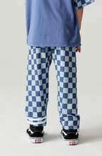 Load image into Gallery viewer, A person shown from the back wearing loose blue and white checkerboard pants, a blue T-shirt, and black Vans sneakers against a plain light studio background.
