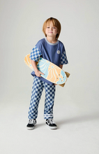 Load image into Gallery viewer, A young child stands against a light gray studio background holding a skateboard. The child is wearing a blue short-sleeve T-shirt with blue-and-white checkerboard sleeves, matching blue-and-white checkerboard pants, and black sneakers.

