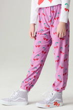 Load image into Gallery viewer, A child wearing pink jogger pants with a red cherry print, a white long-sleeve top with a red heart, and white sneakers, shown from the waist down against a light gray studio background.

