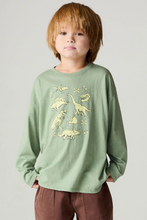 Load image into Gallery viewer, A young boy with light brown hair wearing a sage green long-sleeve t-shirt with a dinosaur print, paired with brown pants, standing against a light gray background.
