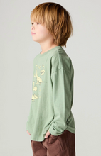 Load image into Gallery viewer, A child model in profile wearing a sage green long-sleeve t-shirt with a dinosaur graphic print on the front, paired with brown pants, against a light gray studio background.
