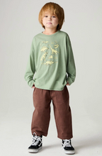 Load image into Gallery viewer, A young child modeling a sage green long-sleeve shirt with a yellow dinosaur graphic, paired with brown pants and black sneakers, against a light gray studio background.
