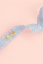 Load image into Gallery viewer, Pair of gold-tone snowflake stud earrings resting on a pale blue ribbon against a peach background.⠋
