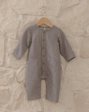 Load image into Gallery viewer, A baby long-sleeve romper in a brown and beige gingham check pattern, hanging on a wooden hanger against a light stone wall.
