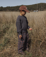 Load image into Gallery viewer, A child standing in a dry grassy field wearing a dark gray cable-knit sweater, plaid pants, and a brown cap.

