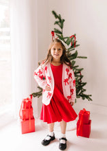 Load image into Gallery viewer, Full-length front view of a child wearing the blush pink bow cardigan over a red dress beside a Christmas tree.
