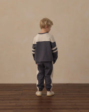 Load image into Gallery viewer, A young boy stands with his back to the camera wearing a navy blue and cream color-blocked sweatshirt and matching sweatpants, standing on a dark wood floor against a plain beige wall.
