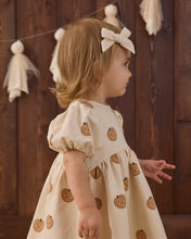 Load image into Gallery viewer, A toddler girl in profile wearing a cream-colored dress with a pumpkin print and a matching cream bow headband, standing in front of a dark wood paneled wall with decorative tassels.
