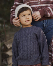 Load image into Gallery viewer, A young child wearing a dark blue-gray cable-knit sweater and a cream knit beanie, standing outdoors while an adult in a brown and cream patterned sweater adjusts the beanie.
