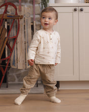 Load image into Gallery viewer, Toddler looking upward while wearing a cream rocking-horse print shirt with tan pants beside wooden sleds.
