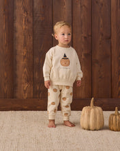 Load image into Gallery viewer, A toddler stands barefoot in front of a dark wood plank wall, wearing a cream two-piece pajama set with a pumpkin graphic on the sweatshirt and pumpkin prints on the pants. Two small pumpkins sit on the woven rug beside the child.
