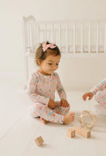 Load image into Gallery viewer, Baby sitting cross-legged beside wooden toys in pale pink Sugar Plum zip pajamas with nutcracker and sweets motifs.
