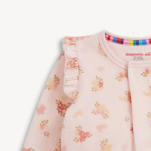 Load image into Gallery viewer, Close-up of a baby long-sleeve top in light pink fleece with a floral print, ruffle detail on the shoulder, and a colorful striped inner neck trim. The brand 'magnetic me' is visible on the inside neck label.

