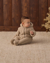 Load image into Gallery viewer, Baby looking to the side in a latte tan fleece footed jumpsuit with the hood down, showing the front zipper.
