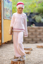 Load image into Gallery viewer, A child wearing a fuzzy light pink two-piece knit outfit with red trim, a matching pink and red beanie, and gray sneakers, standing on a tree stump in an outdoor play area.

