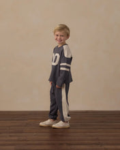 Load image into Gallery viewer, A young boy stands on a dark wood floor against a textured beige wall, wearing a matching navy and cream athletic tracksuit with white sneakers.
