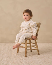 Load image into Gallery viewer, A toddler sits on a small rustic wooden chair wearing a cream ruffled long-sleeve top and matching pants with tiny dark blue star prints, set against a warm beige textured wall and rug.
