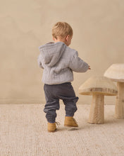 Load image into Gallery viewer, A toddler seen from behind wearing a gray and white gingham patterned hooded jacket, dark gray pants, and tan boots, standing on a textured cream rug next to wooden mushroom-shaped stools.
