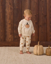 Load image into Gallery viewer, A toddler stands barefoot on a woven rug in front of a dark wood plank wall, wearing a cream-colored pajama set with a pumpkin graphic on the sweatshirt and small pumpkin prints on the pants. Two decorative pumpkins sit on the floor to the right.
