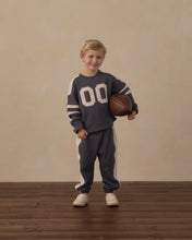 Load image into Gallery viewer, A young boy stands on a wooden floor against a beige wall, wearing a dark gray and white varsity-style sweatshirt and sweatpants set while holding a brown basketball.
