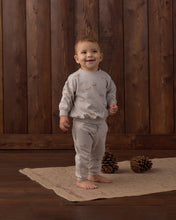 Load image into Gallery viewer, A smiling toddler stands barefoot on a woven rug in front of a dark wood plank wall, wearing a light gray long-sleeve pajama top and matching pants with a small brown animal print. Two pinecones sit on the rug beside the child.
