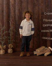 Load image into Gallery viewer, A smiling young boy stands in front of a rustic dark wood plank wall, wearing a cream long-sleeve polo shirt with navy sleeve stripes and the number 14 on the chest, dark pants, and brown boots. Decorative wood signs and bundles of dried grass sit on either side.
