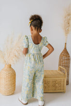 Load image into Gallery viewer, Child wearing a floral jumpsuit standing in front of decorative vases and a woven box.
