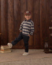 Load image into Gallery viewer, A young child wearing a navy and gray striped long-sleeve polo shirt, dark pants, white sneakers, and a brown cap, posing in front of a rustic dark wood plank wall.

