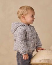 Load image into Gallery viewer, A toddler boy wearing a gray and white gingham patterned hooded puffer jacket and dark gray pants, standing in profile against a warm beige textured wall.
