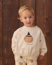 Load image into Gallery viewer, A toddler wearing a cream-colored sweatshirt and matching pants set with a cute pumpkin graphic, standing against a dark wood plank background.
