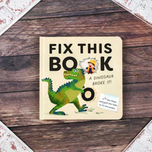 Load image into Gallery viewer, A children's board book titled 'FIX THIS BOOK: A DINOSAUR BROKE IT!' lies flat on a dark wood surface. The cover is cream-colored with bold black text, a green dinosaur illustration, and a small torn-paper detail showing a hand holding a broken tooth.
