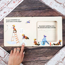 Load image into Gallery viewer, An open children's book with illustrated construction-themed pages, placed on a dark wood surface with a hand pointing at the left page. The book shows cartoon workers, a ladder, scissors, paint, and text including 'He just CHOMPED two giant holes on the page!' and 'Good job, building crew.
