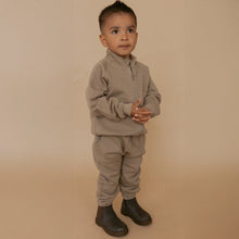 Load image into Gallery viewer, A toddler boy wearing a matching taupe fleece zip-up jacket and sweatpants set, paired with dark brown boots, standing against a solid beige studio backdrop.
